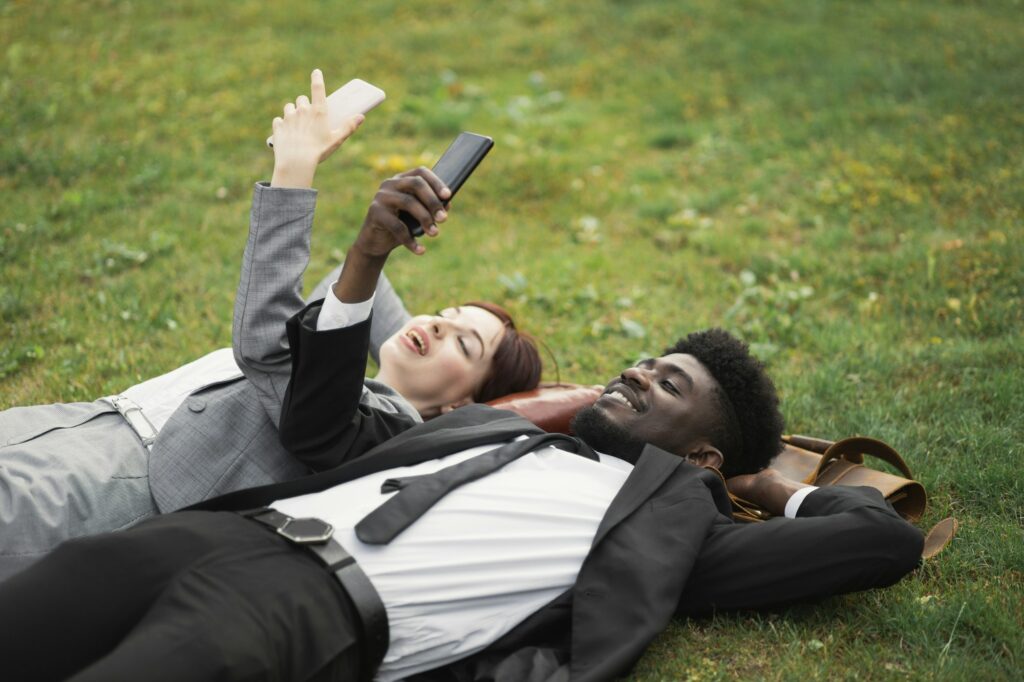 Multiracial well-dressed young business people resting lying on green grass with smartphone